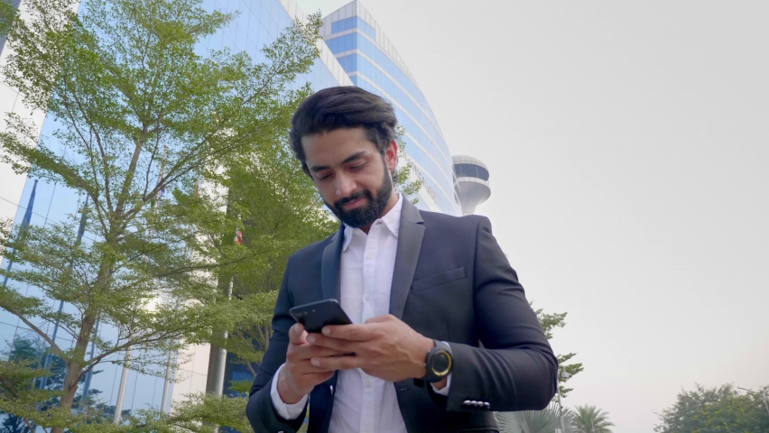 A happy bearded Indian office executive businessman walks by a corporate building using a mobile phone to type a text message. smiling Asian handsome male employee outside a modern commercial complex Royalty-Free Stock Footage #1085817710