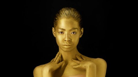 Female With Golden Skin Touching Neckline And Stroking Perfect Golden Body Standing Posing Over Black Background, Studio Shot. Bodycare And Fashion Art Concept