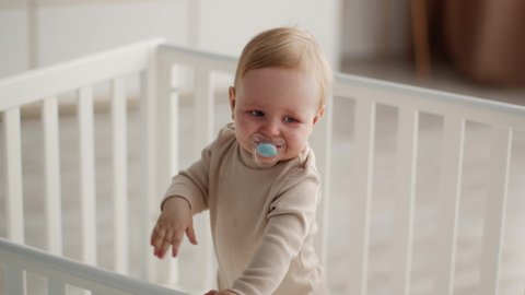 Adorable Toddler Baby With Pacifier In Mouth Standing In Crib And Waving Hand, Closeup Portrait Of Cute Infant Boy Or Girl Using Soother Playing In Bed, Having Fun At Home, Slow Motion Footage