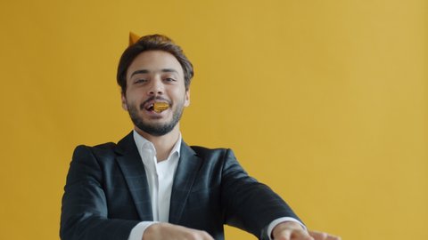 Slow motion portrait of Middle Eastern man wearing party hat blowing whistle celebrating birthday having fun alone on yellow background