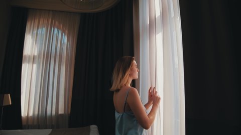 Young happy woman in elegant sleepwear standing by windows to open dark curtains and admire city views. Cityscape in glass window. People in home.