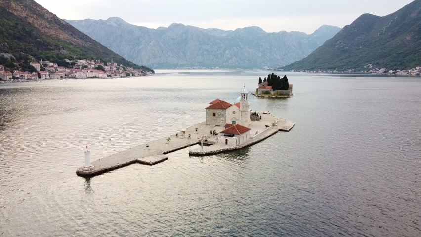Fly over two small island with buildings in a bay. Mountains in the background. Drone view. Drone Shot of Our Lady of the Rocks in Montenegro.