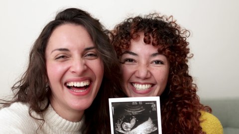 POV LGBT lesbian couple doing video call holding ultrasound photo scan of growing baby in pregnancy time