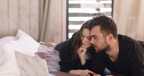 Happy millennial man and woman using laptop computer at home watching online show lying in bed together. Young adult couple looking at notebook screen watch movie or film in bedroom at home.