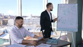 Two businessmen discussing something in a light office. One man sitting at the table and holding a paper. Business partner standing at the whiteboard looking at the chart and commenting it. - Powered by Shutterstock - Get 15% off with code: PIKWIZARD15