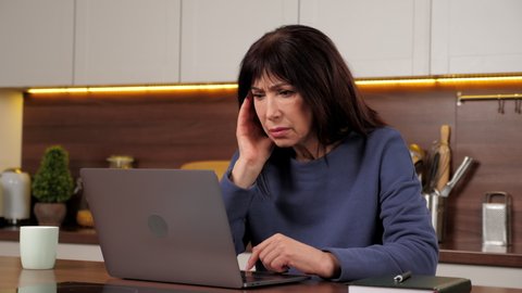 Desperate businesswoman uses laptop reviews company sales report feeling worried about financial problem. Stressed woman works for computer frustrated thinking of money debt, sitting in home kitchen