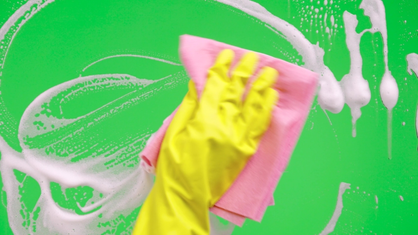 Woman's hand in rubber yellow glove washes window on green screen chroma key background. Housekeeper wipes soap suds from the glass. Cleaning windows with cleaning disinfectant. Close up. Royalty-Free Stock Footage #1085846060