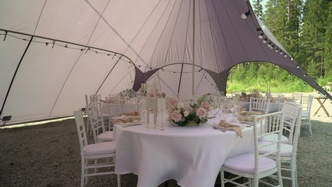 Served table in a tent for dinner, lunch or celebration. White tablecloth, plates, napkins and glasses, chairs. Holiday pavilion. Empty with no people and food. Decorated with flowers for wedding.