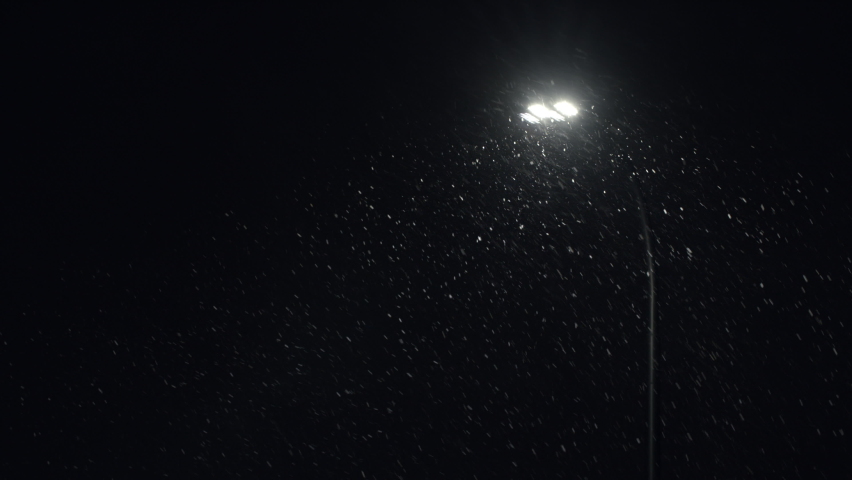Falling snow backlit with modern street lamp in city street at night in winter. Snowflakes illuminated by bright light in park. Snowfall, snow storm and and strong gusty wind Royalty-Free Stock Footage #1085849603