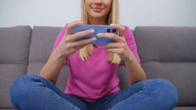 Playing mobile games on smartphone. Happy blonde female plays video game on modern mobile phone while sitting on couch at home. Cheerful Caucasian person with blonde hair using phone for entertainment