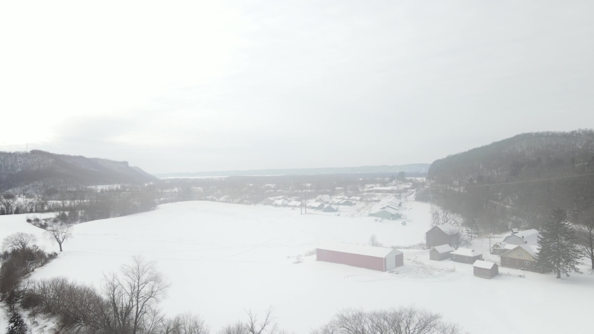 Snow falling in Wisconsin valley in winter. Wide open field with homes seen in the distance. Mountains on each side with gray cloudy sky. 