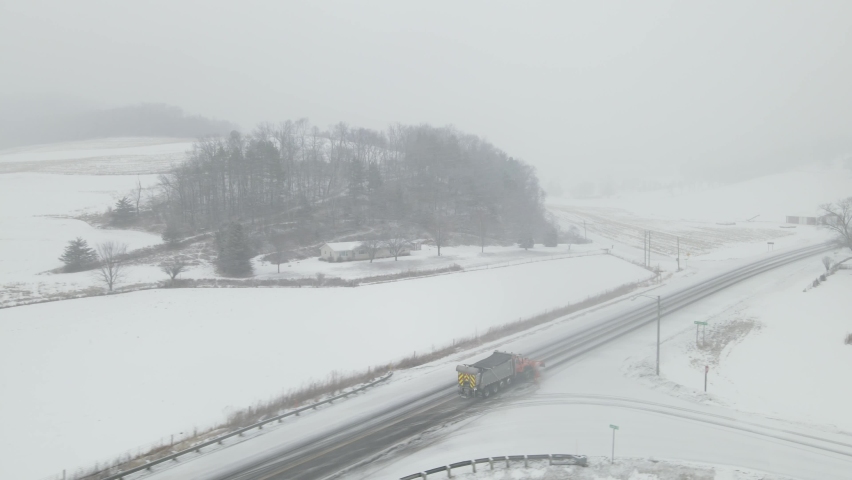 Aerial view following snow plow down snow covered rural highway. Farm land covered with snow. Background obscured by hazy snowy sky. 