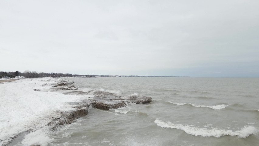 Waves from Lake Michigan splashing against the rough shoreline and freezing on the ground. Water tower visible behind building. Forest seen farther down the shoreline. 