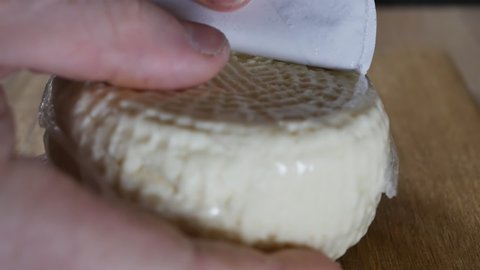human hands unpacked a hard round loaf of vacuum-packed cheese and placed it on a wooden board. Fresh cheese for frying