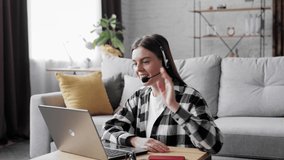 Young caucasian woman wearing wireless headset making conference call working from home sitting on sofa at home. Female distant online teacher doing video chat communicating by webcam on laptop