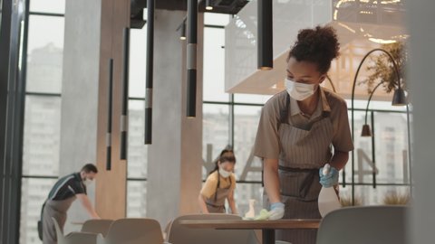 Low angle of young Biracial female cafe employee wearing face mask and gloves, wiping table with disinfecting liquid in modern restaurant, her colleagues working on background