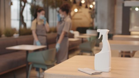 Steadicam of white sprayer bottle and towel on table in restaurant, two blurred workers standing on background, greeting