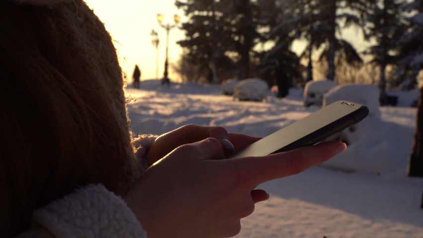 Close-up hands of unrecognizable young woman using mobile phone on winter snowy city street on background of sunlight sunset. Tracking shot of female typing online message in sunny winter day.