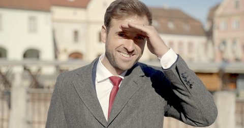 close up of happy man in elegant suit holding hand to forehead to protect eyes and sight from sun while looking far away in the city