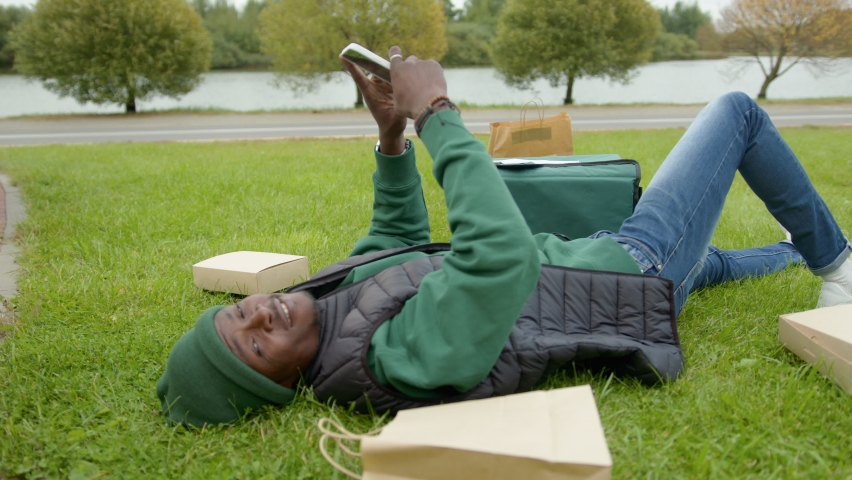 Young black delivery man lies on the lawn in the city park next to the boxes