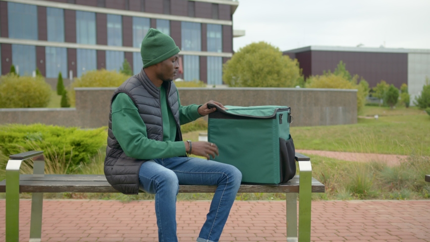 A young black delivery man in a city park on a bench.