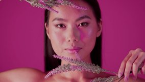 Korean Cosmetics Concept. Beautiful Asian Female Holding Flowers Near Face And Looking At Camera While Posing Over Pink Studio Background, Attractive Young Woman Enjoying Natural Beauty, Slow Motion
