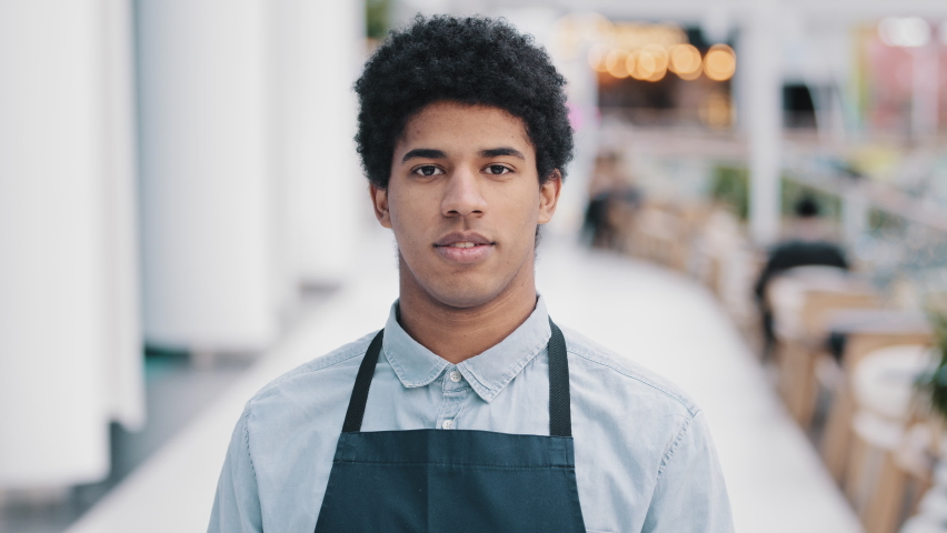 Friendly african american young guy male waiter salesman restaurant cafe kitchen bar worker in apron looking at camera showing gesture her you come here approach welcome inviting customers promotions Royalty-Free Stock Footage #1085871044