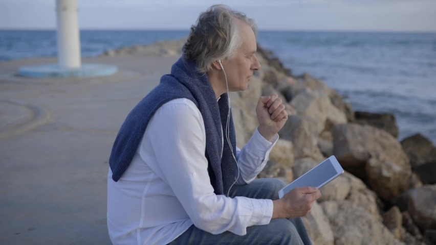 Mature man sitting at beach listening music from digital tablet