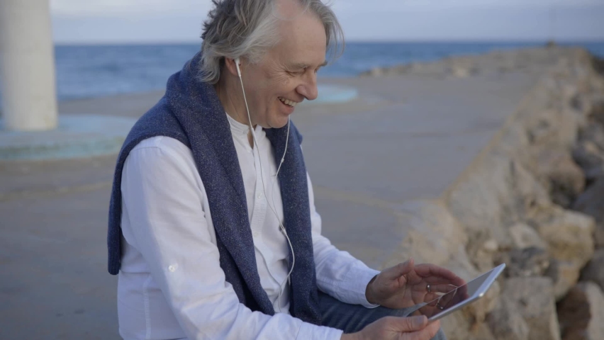 Mature man sitting at beach listening music from digital tablet