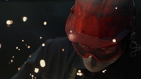 Industrial Welder in the Protective Mask and Orange Glasses and Bright Red Helmet Working With Metal, Dark Background and Sparks comes out from angle grinder, Close Up, Slow Motion