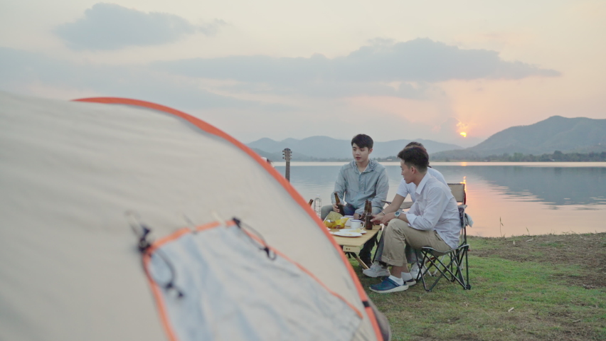 Group Four People Friends Asian men and women Camping, drinking beer, celebrating, Singing, Playing Guitar, Having fun and Enjoying Ground tent. Reservoir area during Sunset Vacation Time. Royalty-Free Stock Footage #1085890682