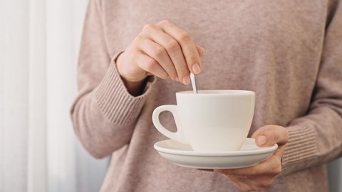 Hands of young woman girl in cozy beige sweater stirs a spoon in cup of tea or coffee near the window. Hands with gentle manicure hold cup with saucer close-up