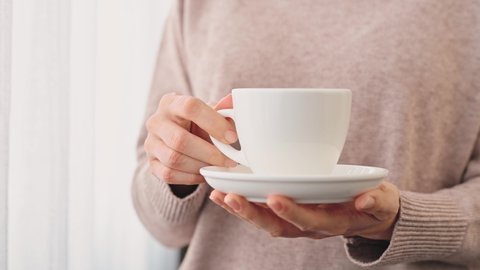 Hands of young woman girl in cozy beige sweater hold white ceramic cup of hot tea or coffee with a saucer near the window. Hands with gentle manicure hold cup close-up