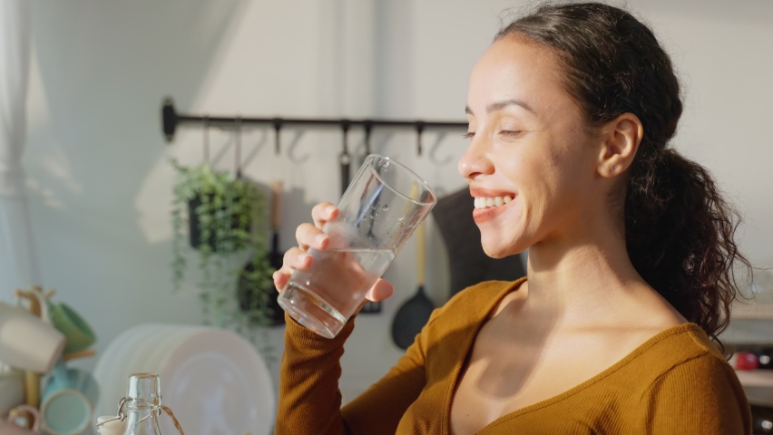 Young beautiful Latino woman drink a glass of clean water in kitchen. Attractive active thirsty girl pouring mineral natural in cup and take a sips for health care and wellbeing in kitchen in house. | Shutterstock HD Video #1085910917