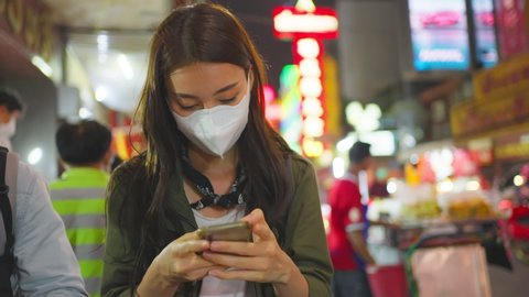 Asian attractive woman backpacker chatting on phone while walk in city. Beautiful woman tourist traveler wear mask while travel alone on street, using smartphone communicate with friend at night road.