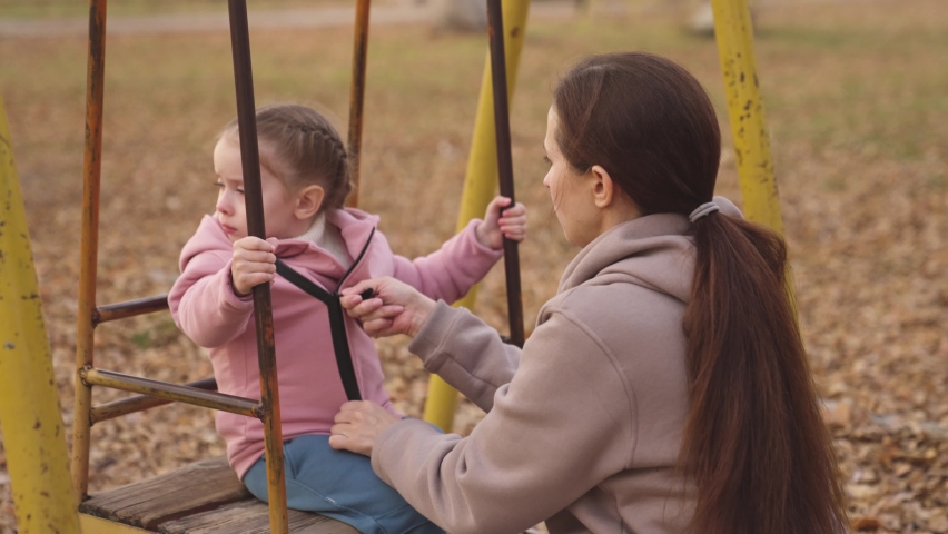 Mom fastens lock on blouse for little daughter in an autumn park, a mother dresses her daughter on playground, riding children swing in yellow leaves, happy family life, parental care and upbringing Royalty-Free Stock Footage #1085911460