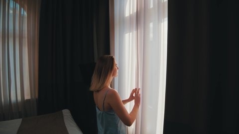 Young dreamy woman in sleepwear opening curtains of window in cozy bedroom. Elegant caucasian girl in apartment enjoying cityscape views and sunshine.