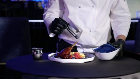 A professional chef in black gloves decorates a dish of fried fruits and citrus fruits, duck confit in the restaurant hall.Cooking in front of customers. Exquisite French haute cuisine