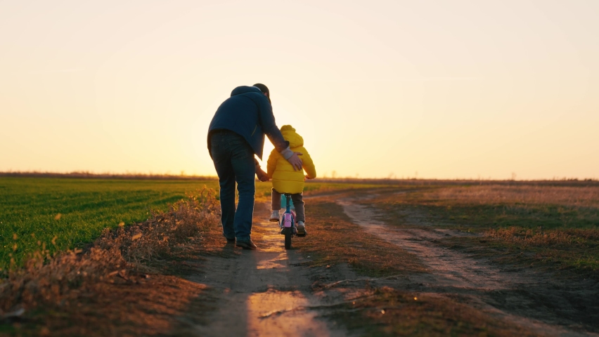 Dad teaches a little girl to ride a children's bike on the road in spring. Happy family. A father teaches his daughter to ride a bike in the park at sunset. Daddy and kid play together outdoors Royalty-Free Stock Footage #1085914925