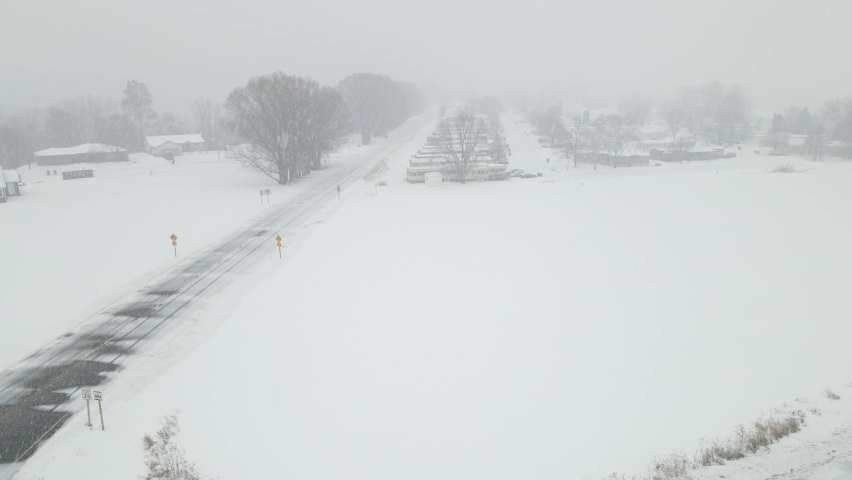 Snow filled sky in a valley in Wisconsin. Mobile home park tucked into the trees. Country road with wind blown snow covering. Gray hazy sky. 