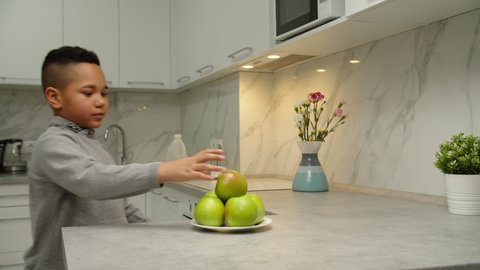 Moving shot of bunch of green apples while carefree black boy approaching kitchen counter, taking fruit, biting off apple, smiling leaning on table indoors. Hungry kid having healthy snack at home