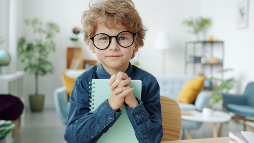 Slow motion portrait of adorable child wearing eyeglasses holding book smiling and looking at camera in apartment. Childhood and education concept. Royalty-Free Stock Footage #1085939618