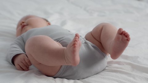 Childbirth Concept. Closeup Shot Of Chubby Newborn Baby Wearing Diaper And Bodysuit Lying On Bed At Home, Small Infant Child Relaxing In Bedroom, Selective Focus On His Tiny Feet, Slow Motion Footage