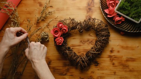 Female hands decorates heart shaped floral wreath. DIY spring home decor crafts. Woman makes Valentines day home decoration with dried twigs and diy flowers. Table top view, POV.