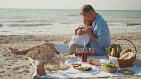 loving young couple happy man and woman sits on cozy picnic blanket with wine, fruits, basket with bread. beautiful couple having romantic date at sea beach, hugging and kissing. happy valentines day.