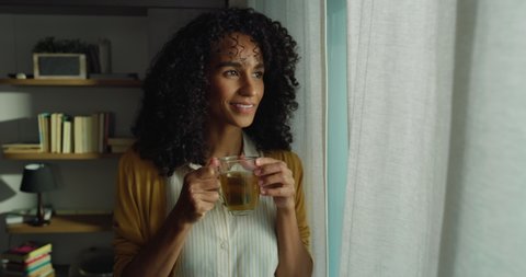 Cinematic shot of young serene smiling brazilian woman is drinking hot tea while looking through window with sunshine at home.