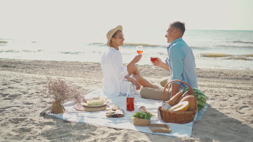 Happy young attractive couple celebrating with a glass of wine on the beach at sunny morning. man and woman in white wear and hats having picnic at sea beach Royalty-Free Stock Footage #1085947808
