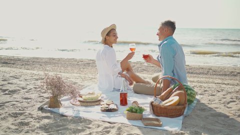 Happy young attractive couple celebrating with a glass of wine on the beach at sunny morning. man and woman in white wear and hats having picnic at sea beach