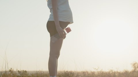 Pregnant white woman stands against background sunset and drinks water from bottle, closeup. Silhouette of pregnant woman, side view.