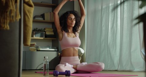 Cinematic shot of young sporty brazilian woman wearing bra and leggings is practicing yoga and meditation seated on mat while relaxing her mind at home